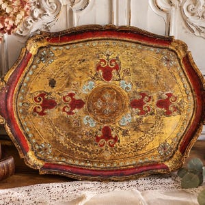 May include: An oval-shaped decorative tray featuring a distressed gold finish with deep red borders and intricate light blue floral accents. The tray has a scalloped edge and integrated handles on both sides, displaying a weathered, aged texture.