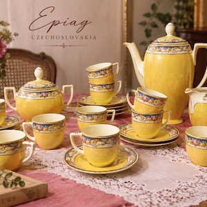 May include: A yellow porcelain tea set by Epiag Czechoslovakia featuring a floral pattern with blue and gold accents. The set includes a teapot, a lidded sugar bowl, a creamer, and multiple teacups with matching saucers, all arranged on a pink cloth.