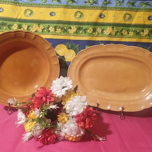 Puede incluir: Dos platos de servir de cerámica, uno redondo y otro ovalado, ambos de color marrón claro. Los platos están sobre una superficie rosa con un arreglo floral en el centro.