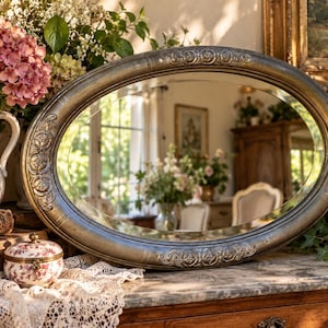 May include: An oval mirror with a dark, weathered metallic frame featuring embossed floral and scrollwork details. The reflective glass surface shows a room interior, while the frame displays a textured, aged finish with visible patina.