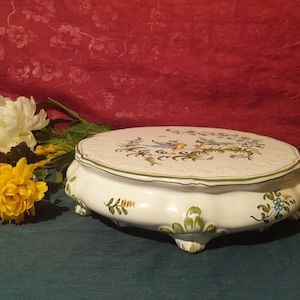 Può includere: Un porta torta in ceramica bianca con un design floreale verde e blu. Il supporto ha tre gambe e un bordo ondulato.