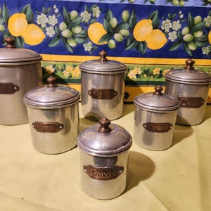Puede incluir: Seis botes de metal plateado con tapas y etiquetas de cobre. Las etiquetas dicen "Epices", "Poivre", "The", "Farine", "Cafe" y "Sucre".