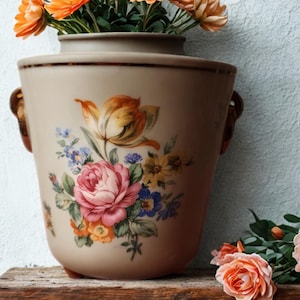 May include: A beige ceramic vase with a floral design featuring pink roses, yellow tulips, and blue wildflowers. The vase has two brown handles and is filled with orange flowers. The vase is sitting on a wooden surface.