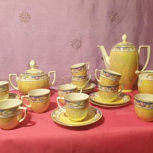 May include: A complete vintage tea set in yellow with floral detailing. The set includes a teapot, sugar bowl, creamer, cups, and saucers. The cups and saucers are arranged on a red surface, with a pink floral backdrop.