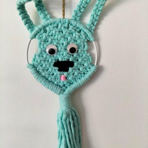 May include: A turquoise macrame bunny wall hanging with googly eyes, a black nose, and a pink tongue. The bunny has long ears and a tassel at the bottom. A gold chain is attached for hanging.