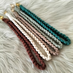 May include: A set of five macrame wristlet lanyards in dark brown, cream, tan, sage green, and dark teal. Each braided cord features a gold-toned metal lobster clasp at the top, showcasing a consistent knotted design and a looped structure.