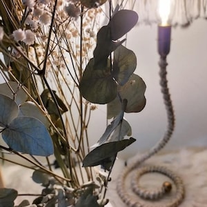 Peut inclure: Un arrangement décoratif de fleurs séchées et de feuilles d'eucalyptus dans un vase blanc. Une lampe en spirale avec une base violette et une ampoule allumée est en arrière-plan. La tige de la lampe est faite de corde torsadée.
