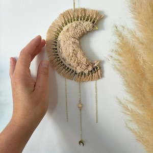 Peut inclure: Décoration murale en forme de croissant de lune en macramé beige avec des accents dorés. La lune présente un centre texturé, des bords frangés et des détails de chaîne dorée avec un charme soleil et lune. Décoration murale sur un mur blanc.