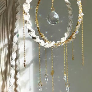 May include: A handcrafted dream catcher with a white macrame outer ring and a gold inner ring adorned with crystals. Gold chains with crystal pendants and charms hang from the rings. A large crystal ball hangs from the center.