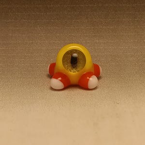 May include: A whimsical, yellow, glass figurine with a single eye and four red and white feet. The eye has a detailed, textured design. The figurine is a unique, decorative item.