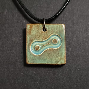 May include: A ceramic pendant necklace with a blue bicycle chain link design on a green and brown background. The necklace has a black cord.