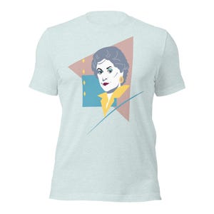 May include: Light blue t-shirt featuring a graphic illustration of a woman's portrait. The portrait is set against a geometric background with a pink triangle, blue square, and yellow accents. The woman has gray hair, red lips, and gold earrings.