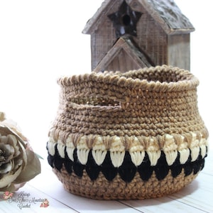 May include: A crocheted basket with a brown, cream, and black color scheme. The basket has a handle and a decorative trim around the top.