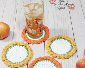 May include: Four handmade crochet coasters with a white centre, bordered by green and orange yarn. A glass of iced tea with pumpkin decals sits on one coaster. Two apples are on the sides. The text "Krissy's Over The Mountain Crochet" is in the top right corner.