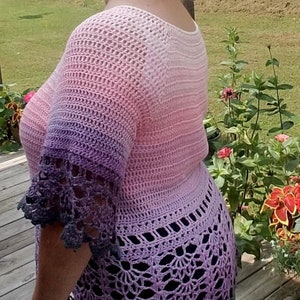 May include: A woman wearing a crocheted top with a gradient of pink, purple, and gray. The top has a lacy trim and is worn over a black skirt. The text "Krissys Over The Mountain Crochet" is visible on the skirt.