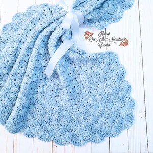 May include: A light blue crocheted baby blanket with a scalloped edge. The blanket is tied with a white satin ribbon. The text "Krissys Over The Mountain Crochet" is visible in the upper right corner of the image.