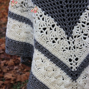 Op de afbeelding: Een grijs en wit gebreid poncho met een kantpatroon. De poncho heeft een V-vormige halslijn en is gemaakt van zacht garen. De tekst "Krissy's Over The Mountain Crochet" is zichtbaar in de linkerbovenhoek van de afbeelding.