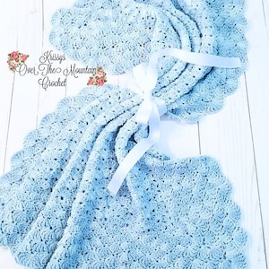 May include: A light blue crocheted baby blanket with a scalloped edge. The blanket is tied with a white satin ribbon. The text "Krissys Over The Mountain Crochet" is visible in the image.