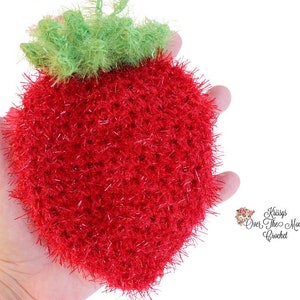 Pode incluir: Uma esponja de cozinha de morango vermelha e verde feita de crochê. A esponja é feita com fio brilhante e é segurada na mão de uma pessoa.