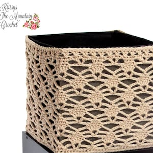 Puede incluir: Una cesta de crochet beige con un forro de tela negro. La cesta tiene forma cuadrada y un delicado diseño calado.