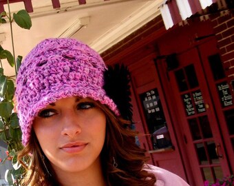 May include: A woman with long brown hair is wearing a pink crocheted hat with a black flower embellishment. She is wearing a pink and white top and has a neutral expression.