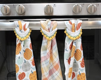 May include: Three kitchen towels with a pumpkin theme hanging on a silver oven rack. The towels are white with a plaid pattern, white with a pumpkin pattern, and white with a pumpkin and leaf pattern. The towels have crocheted loops at the top.
