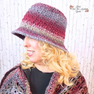 May include: A woman wearing a crocheted hat with a burgundy, gray, and gold color scheme. The hat has a wide brim and is styled in a bucket hat shape. The woman is also wearing a crocheted shawl with a similar color scheme.