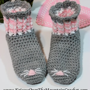 May include: Gray crocheted slippers with pink and white trim and a cat face design. The slippers are on a white surface with a pink doily.