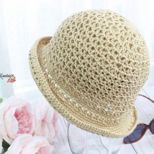 May include: A beige crochet hat featuring an openwork stitch pattern throughout the crown and a structured brim. The hat displays a decorative band with small cutouts above the brim and includes the text Krissys Over The Mountain Crochet.