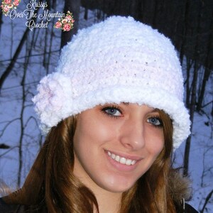 Puede incluir: Un sombrero blanco de ganchillo con una pequeña flor rosa. El sombrero tiene un ala y un aspecto texturizado. El texto "Krissys Over The Mountain Crochet" es visible en la esquina superior izquierda.
