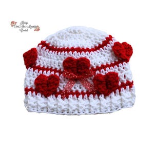 Può includere: Cappello bianco all'uncinetto con strisce rosse e decorazioni a forma di cuore rosso. Il cappello ha un fiocco di nastro rosso con pois bianchi.