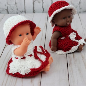 May include: Two crocheted doll dresses with matching hats. One dress is white with red trim and the other is red with white trim.