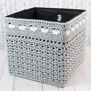 May include: A gray crochet storage basket with a black interior. The basket features a decorative white trim along the top edge. The handmade basket is square-shaped and suitable for organizing items in a home or office.