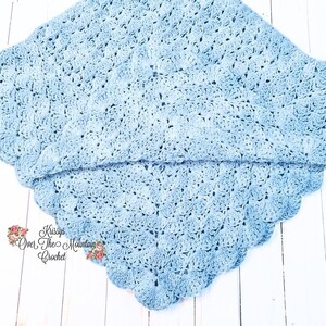May include: A light blue crocheted baby blanket with a scalloped edge. The blanket is folded in half and laid on a white wooden surface. The text "Kissys Over The Mountain Crochet" is visible in the lower right corner of the image.
