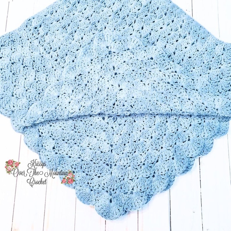 May include: A light blue crocheted baby blanket with a scalloped edge. The blanket is folded in half and laid on a white wooden surface. The text "Kissys Over The Mountain Crochet" is visible in the lower right corner of the image.