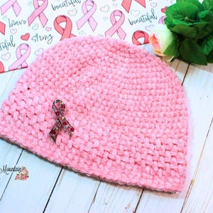 Puede incluir: Un gorro de ganchillo rosa suave con un broche de lazo. El sombrero está adornado con un broche de lazo decorativo. El fondo presenta un papel estampado con lazos rosas y las palabras "Strong" y "Beautiful".