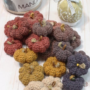 May include: A collection of crocheted pumpkins in various shades of brown, orange, and purple. The pumpkins are arranged on a white wooden surface. The yarn used to crochet the pumpkins is in the background.