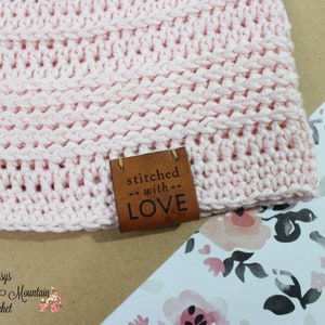 K&ouml;nnte beinhalten: Eine rosa geh&auml;kelte M&uuml;tze mit einem braunen Lederanh&auml;nger, auf dem "stitched with LOVE" steht.