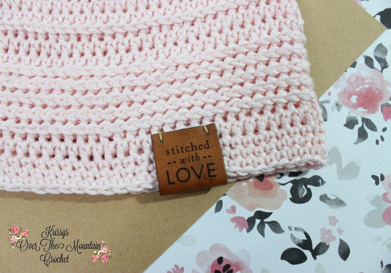 K&ouml;nnte beinhalten: Eine rosa geh&auml;kelte M&uuml;tze mit einem braunen Lederanh&auml;nger, auf dem "stitched with LOVE" steht.