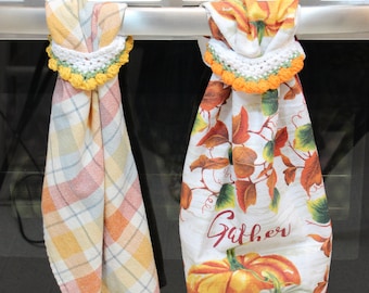 May include: Two kitchen towels hanging on a silver oven handle. The towel on the left is plaid with orange, yellow, and white squares. The towel on the right has a white background with a pumpkin and fall leaves design. Both towels have a crocheted trim with yellow and green accents. The text "Krissys Over The Mountain Crochet" is visible on the oven.