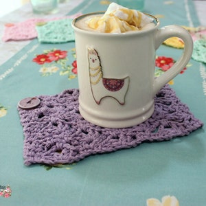 May include: A white ceramic mug with a llama illustration and a gold rim, filled with a creamy beverage topped with whipped cream and caramel sauce. The mug is resting on a purple crocheted coaster.