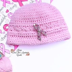 May include: Pink crocheted beanie with a pink ribbon pin. The beanie is made with a textured stitch and has a simple design. The pin is a sparkly, jeweled ribbon.