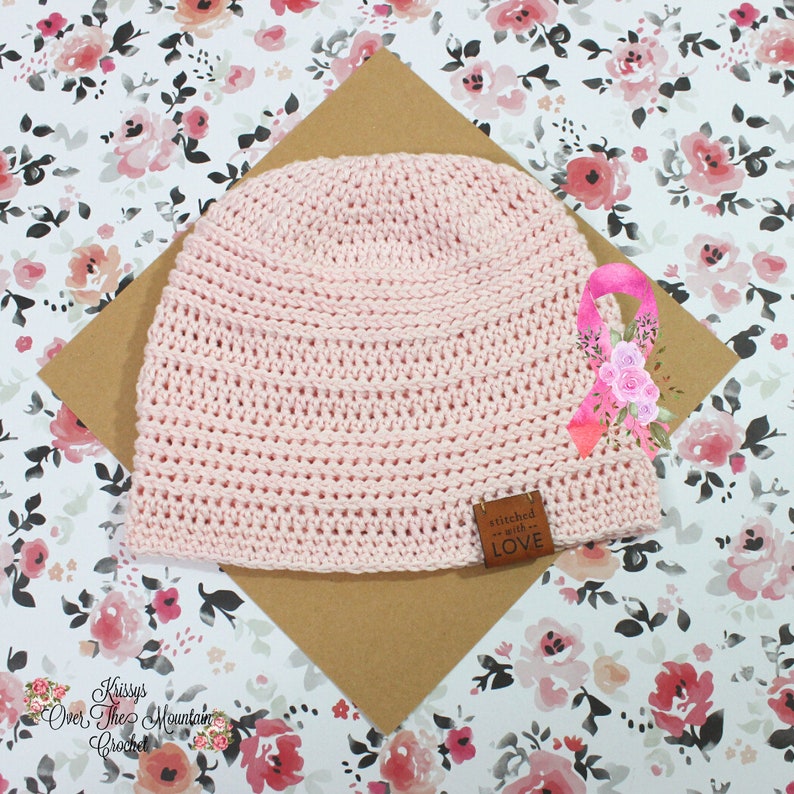 K&ouml;nnte beinhalten: Eine rosa geh&auml;kelte M&uuml;tze mit einem braunen Lederanh&auml;nger, auf dem "mit Liebe gen&auml;ht" steht. Die M&uuml;tze liegt auf einem braunen Hintergrund mit einem rosafarbenen Blumenmuster.