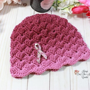 May include: A crocheted beanie in a gradient of dark magenta to light pink featuring a textured shell stitch pattern. A silver-toned awareness ribbon pin with embedded pink crystals is attached to the front center of the hat.