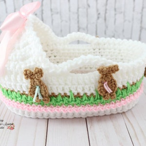 May include: White crocheted baby basket with a pink bow, green and brown trim, and three brown bunny appliques. The basket is on a white wooden surface.
