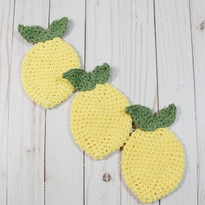 May include: Three crocheted lemon-shaped pot holders with green leaves. The pot holders are yellow and are arranged in a row on a white wooden surface.