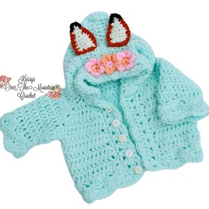 May include: A light blue crochet baby jacket with a hood, featuring fox ears and floral accents. The jacket has a button-up front and scalloped edges. The text "Krissys Over The Mountain Crochet" is visible on the left side.