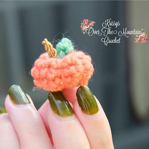 May include: A miniature crocheted pumpkin in orange, green, and gold hues. The pumpkin is held between fingers with green nail polish. The background features the text "Krissys Over The Mountain Crochet".