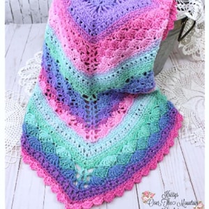 May include: A multi-coloured crochet baby blanket with a scalloped edge. The blanket is made in shades of pink, purple, blue, and green. The blanket is folded and draped over a metal bucket. The text "Over The Mountain Crochet" is visible in the lower right corner of the image.