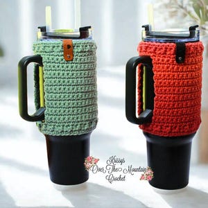 May include: Two insulated tumblers with black handles and lids, each encased in a crocheted cozy. One cozy is sage green, the other is red. Both have a leather strap detail. The tumblers have straws.